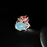 Bague d'artisanat d'art en argent 925 ornée d'une rose sculptée en aigue-marine et d'un papillon en tourmaline rose sur fond rouge bordeaux.