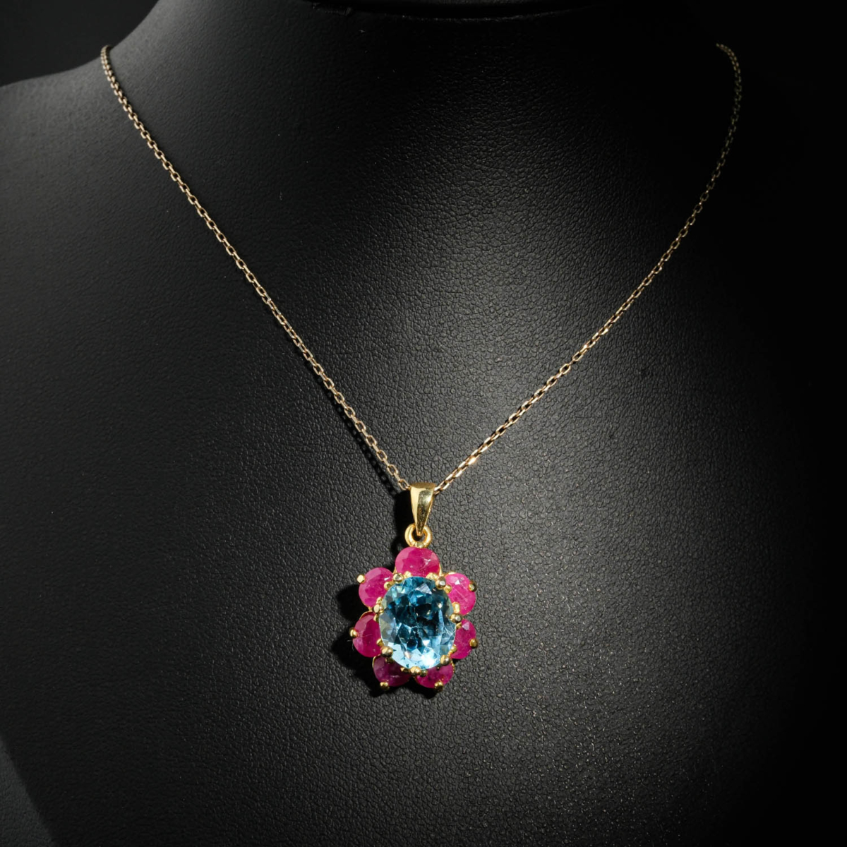 Natural faceted oval blue topaz pendant necklace with ruby flower halo in gold plated 925 silver on white satin.