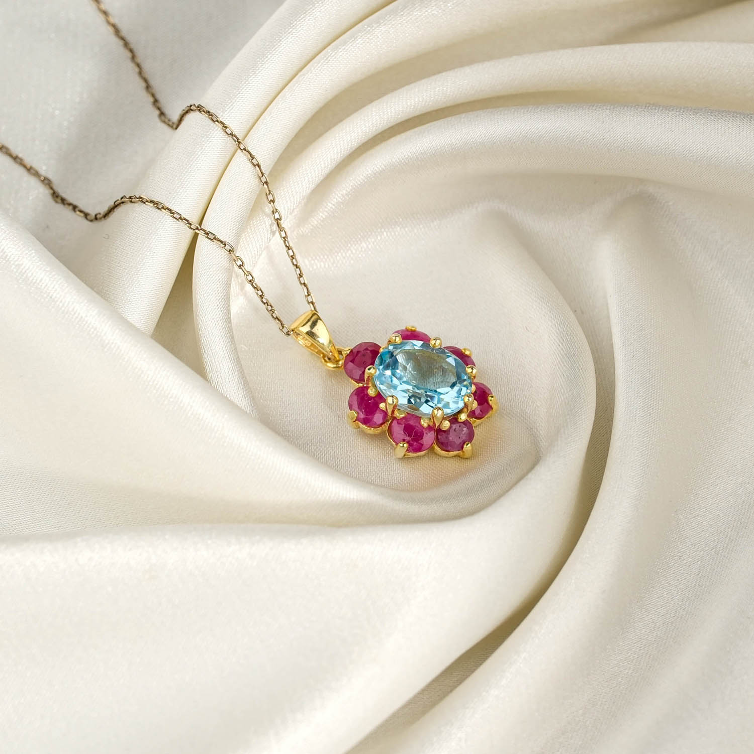 orijinal-blue-topaz-yakut-cicek-925-gumus-gold-kolye.jpg Beyaz saten kumaş üzerinde faset oval mavi topaz etrafında kırmızı yakut taç yapraklı sarı altın kaplama 925 gümüş kadın kolye ucu ve zincir.