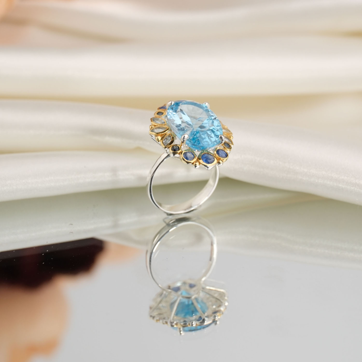 Beyaz saten kumaş ve ayna üzerinde faset oval açık mavi topaz etrafında lacivert safir taşlı altın kaplama hareli 925 gümüş kadın yüzük.