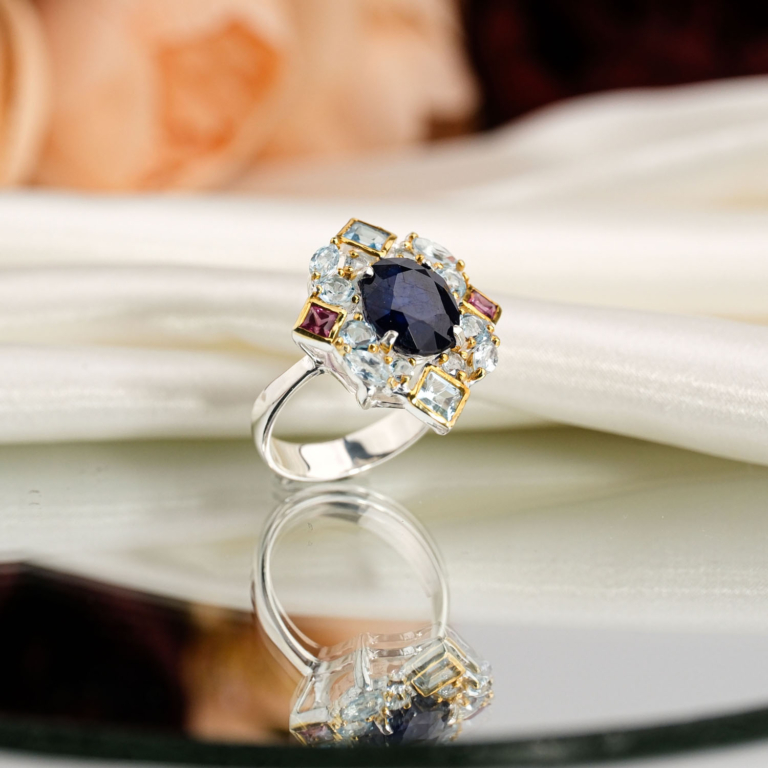 Beyaz saten kumaş ve ayna üzerinde faset oval lacivert safir etrafında açık mavi topaz ve pembe turmalin taşlı altın vurgulu 925 gümüş kadın yüzük.
