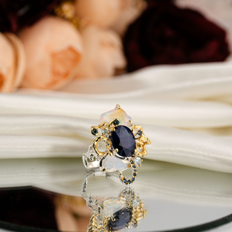 Beyaz saten kumaş ve ayna üzerinde faset oval lacivert safir etrafında yanardöner opal ve sarı altın dallı topaz taşlı 925 gümüş kadın yüzük.