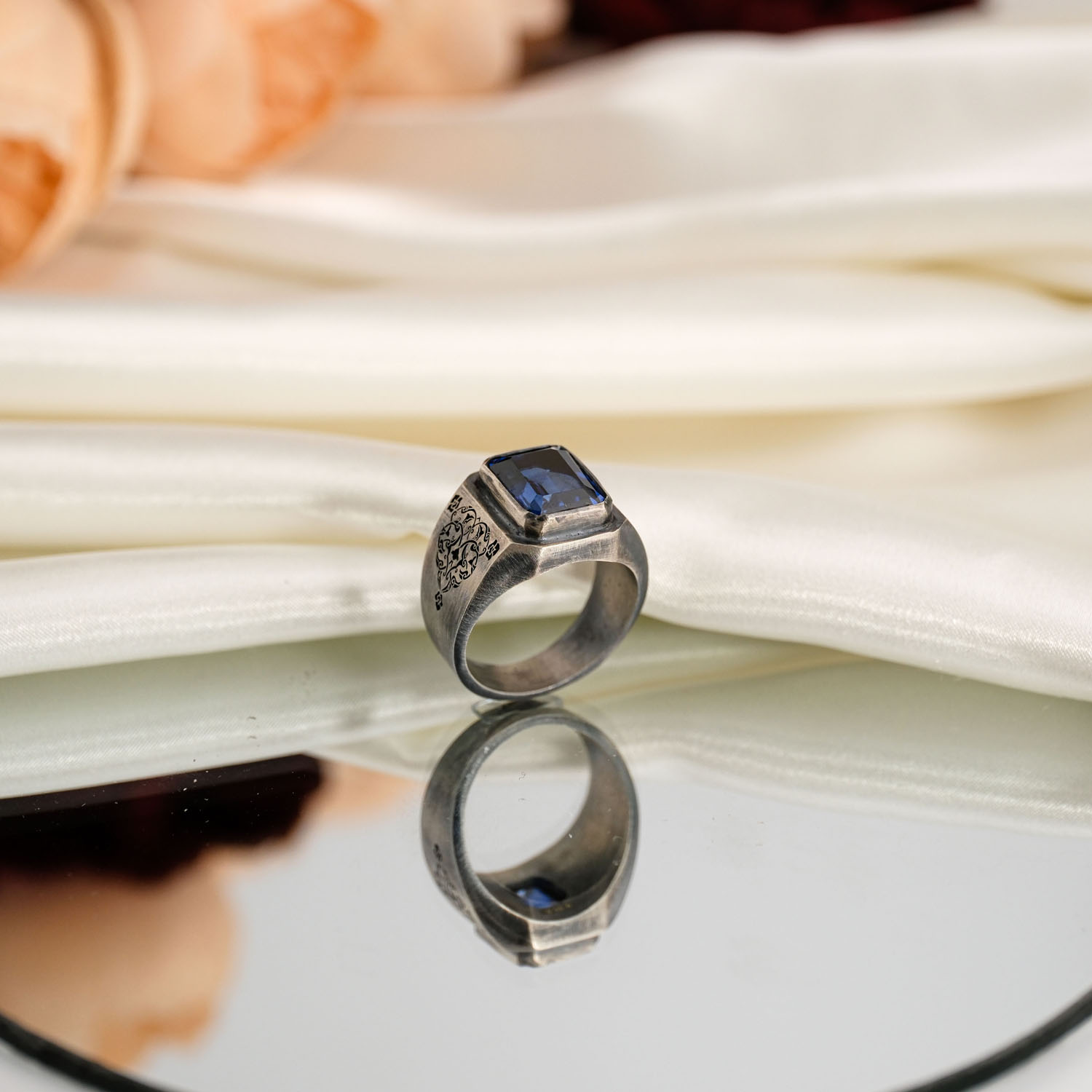 orijinal-safir-5-20ct-baget-el-isi-erkek-gumus-yuzuk.jpg Beyaz saten kumaş ve ayna üzerinde faset baget kesim 5,20 karat koyu mavi safir taşlı kalem işlemeli 925 ayar gümüş erkek yüzük.