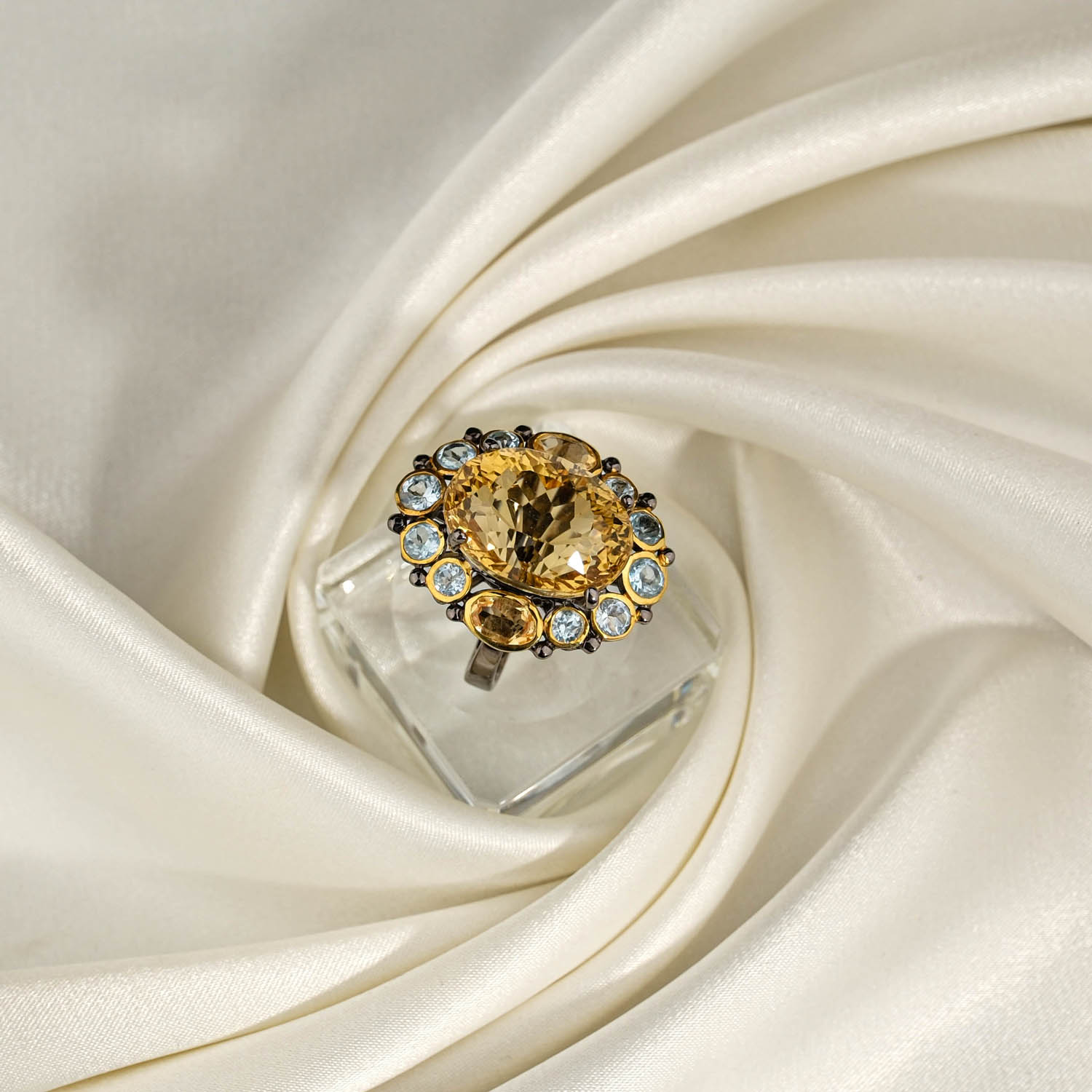orijinal-sitrin-blue-topaz-tasli-925-ayar-gumus-tasarim-yuzuk.jpg Beyaz saten kumaş üzerinde faset oval sarı sitrin taşlı etrafı mavi topaz dizili 925 ayar gümüş tasarım yüzük.